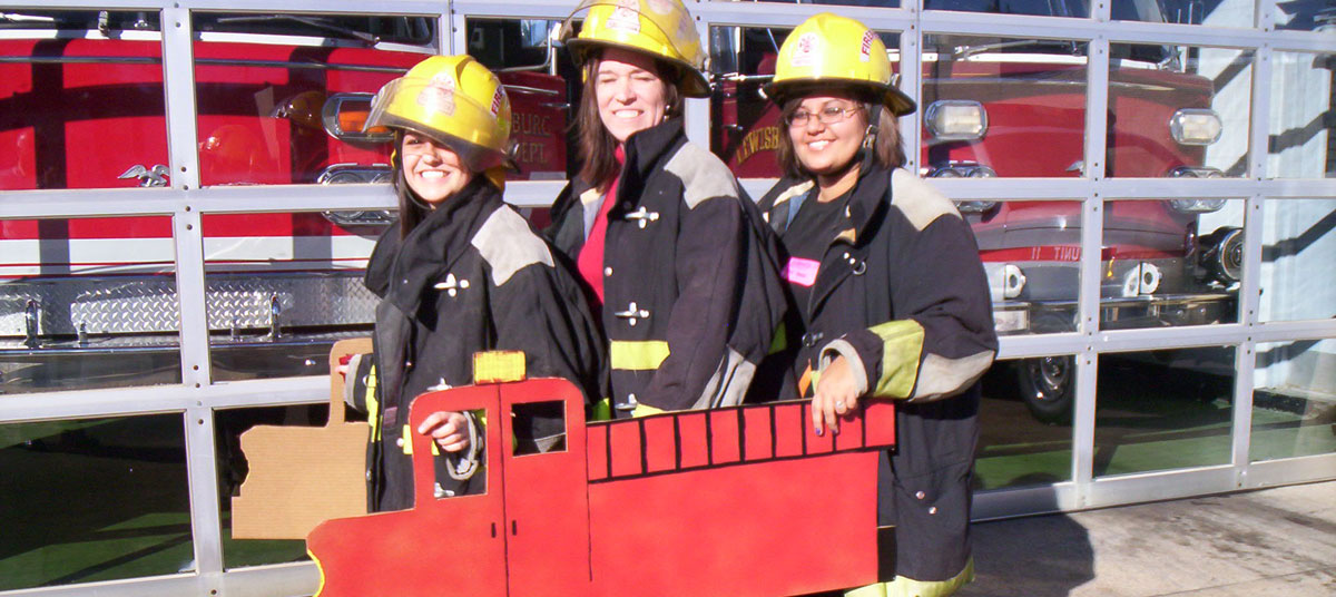 Three girls in firefighter gear holding a cardboard cutout of a fire truck